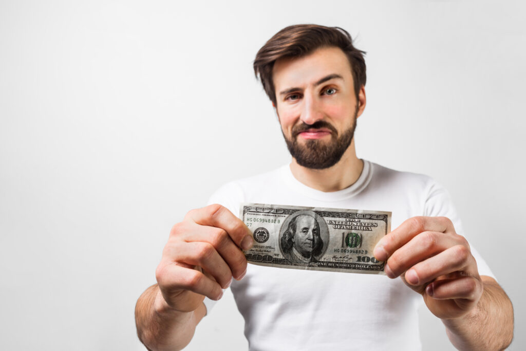 Close up of handsome brunette man standing near the white wall and holding a dollar bill. He is showing that he has one hundred dollar bill. Guy looks happy. Cut view. Isolated on white background.