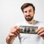 Close up of handsome brunette man standing near the white wall and holding a dollar bill. He is showing that he has one hundred dollar bill. Guy looks happy. Cut view. Isolated on white background.